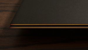 Extreme macro of two razor-thin matte black slab edges stacked on dark walnut, each catching a bright warm amber specular line, illustrating foldable device thickness comparison