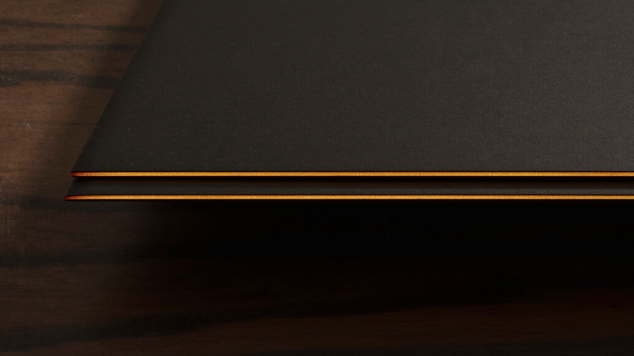 Extreme macro of two razor-thin matte black slab edges stacked on dark walnut, each catching a bright warm amber specular line, illustrating foldable device thickness comparison