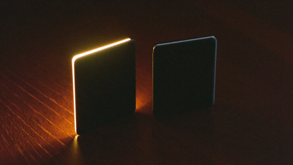 Cinematic editorial hero — two closed foldable phone silhouettes on dark walnut with dramatic warm amber rim light, representing the upcoming Galaxy Z Fold 8 and Z Fold 8 Ultra.