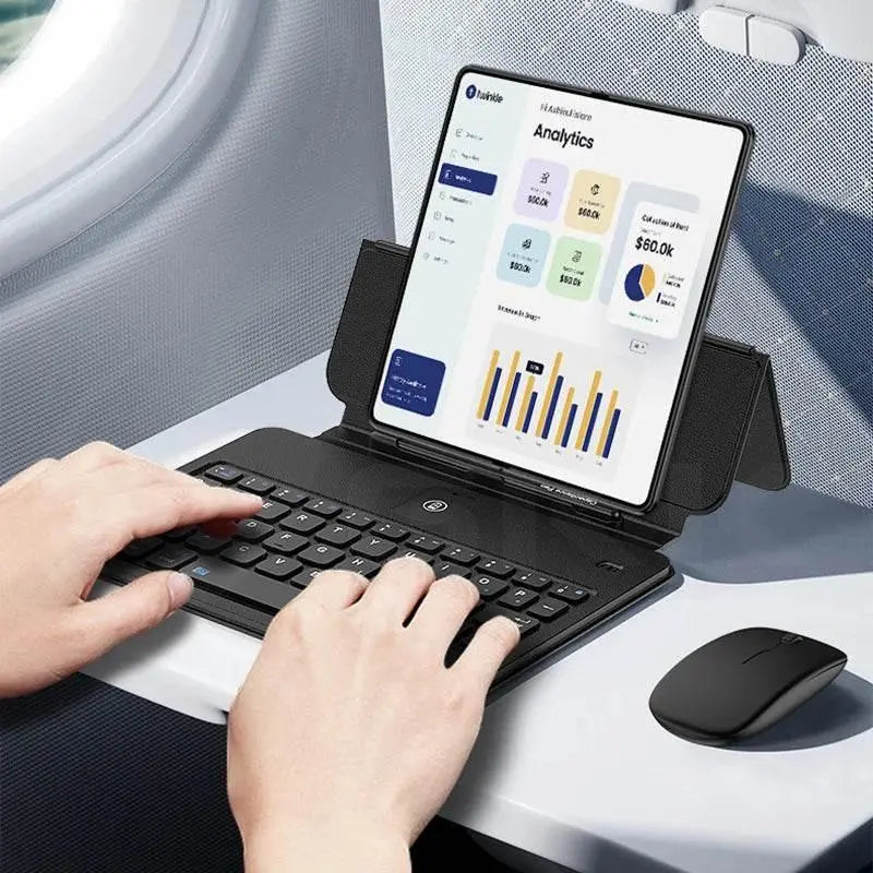 Hands typing on a black foldable Bluetooth keyboard with tablet showing analytics charts on airplane tray table, wireless mouse